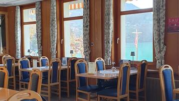 A dining area with wooden tables and blue upholstered chairs, large windows with curtains, and a view of a body of water.
