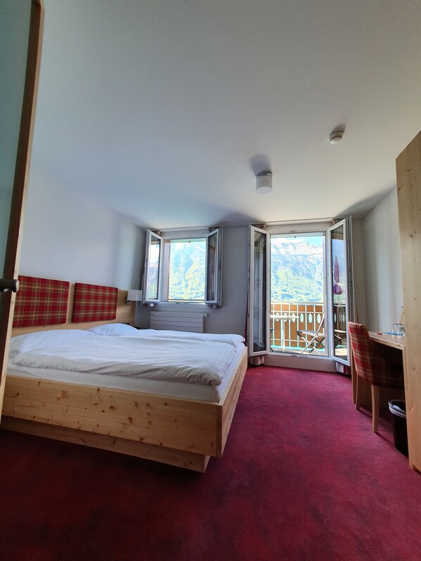 A bedroom with a wooden bed, a desk, and a chair. There are windows with curtains and a view of mountains. - Strandhotel (Iseltwald)