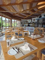 Un restaurante con mesas y sillas de madera, ventanas amplias y una zona de cocina.