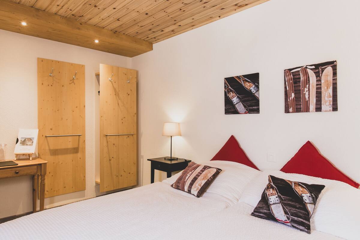 Ein Schlafzimmer mit Holzboden, einem Bett mit weißer Bettwäsche, einem roten Kopfteil und zwei Kissen mit einem Skimotiv. Ein Nachttisch mit Lampe und ein Holztisch mit Stuhl sind ebenfalls vorhanden.