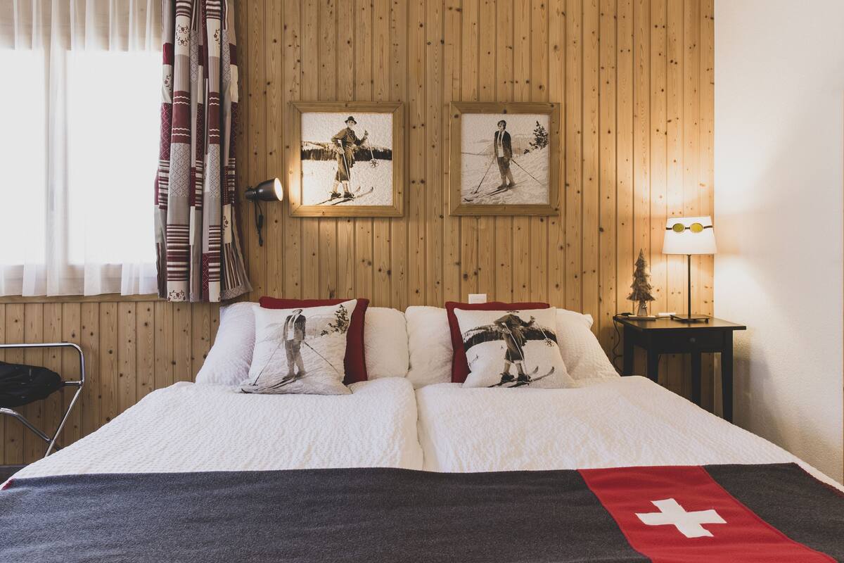 Ein Hotelzimmer mit einer holzgetäfelten Wand, einem Bett mit weißer Bettwäsche und einer roten Decke, zwei gerahmten Bildern von Skifahrern, einer Lampe auf einem Nachttisch und einem Sessel.
