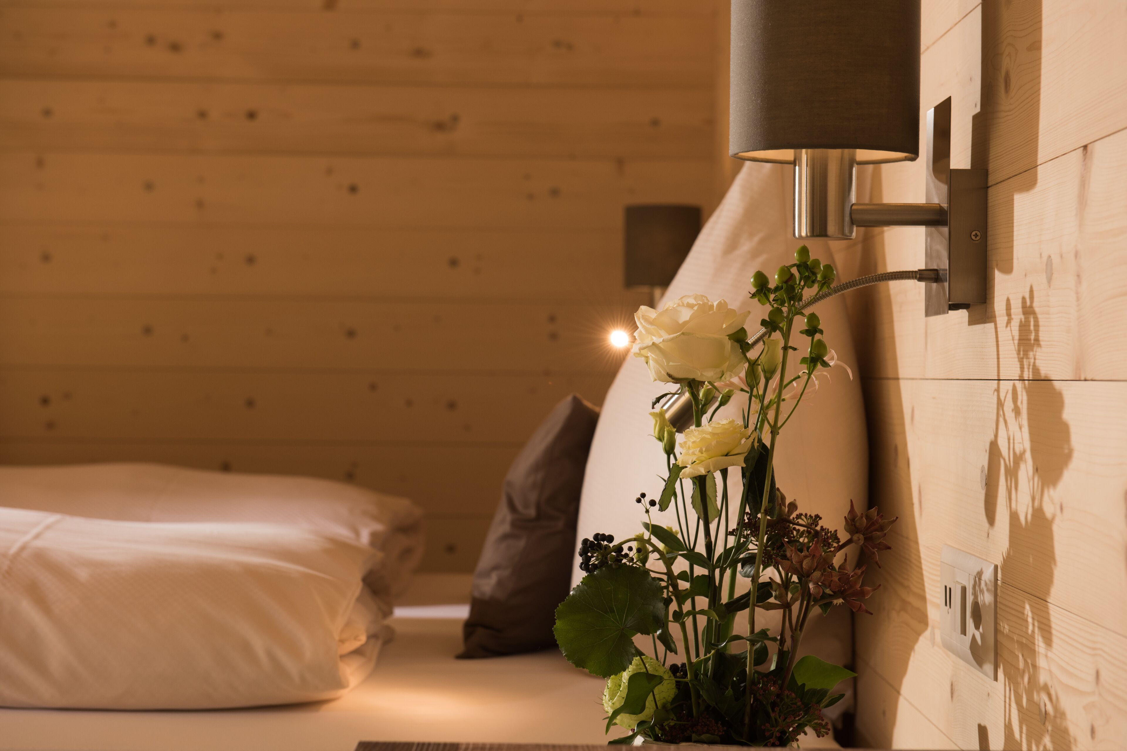 A wooden cabin interior with a bed, pillows, a wall-mounted lamp, and a potted plant with flowers.