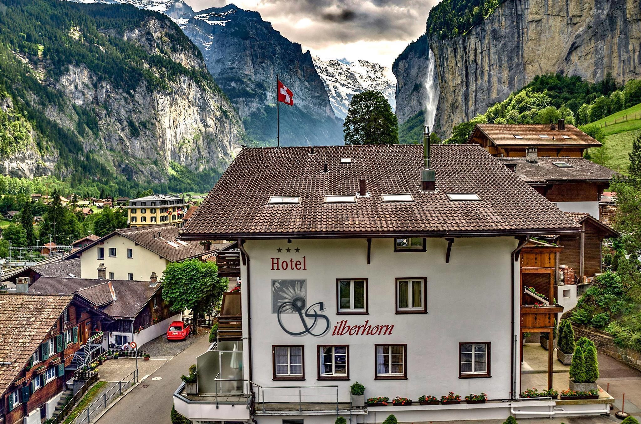 A hotel with a mountain backdrop, featuring a prominent Swiss flag and a sign indicating its name.