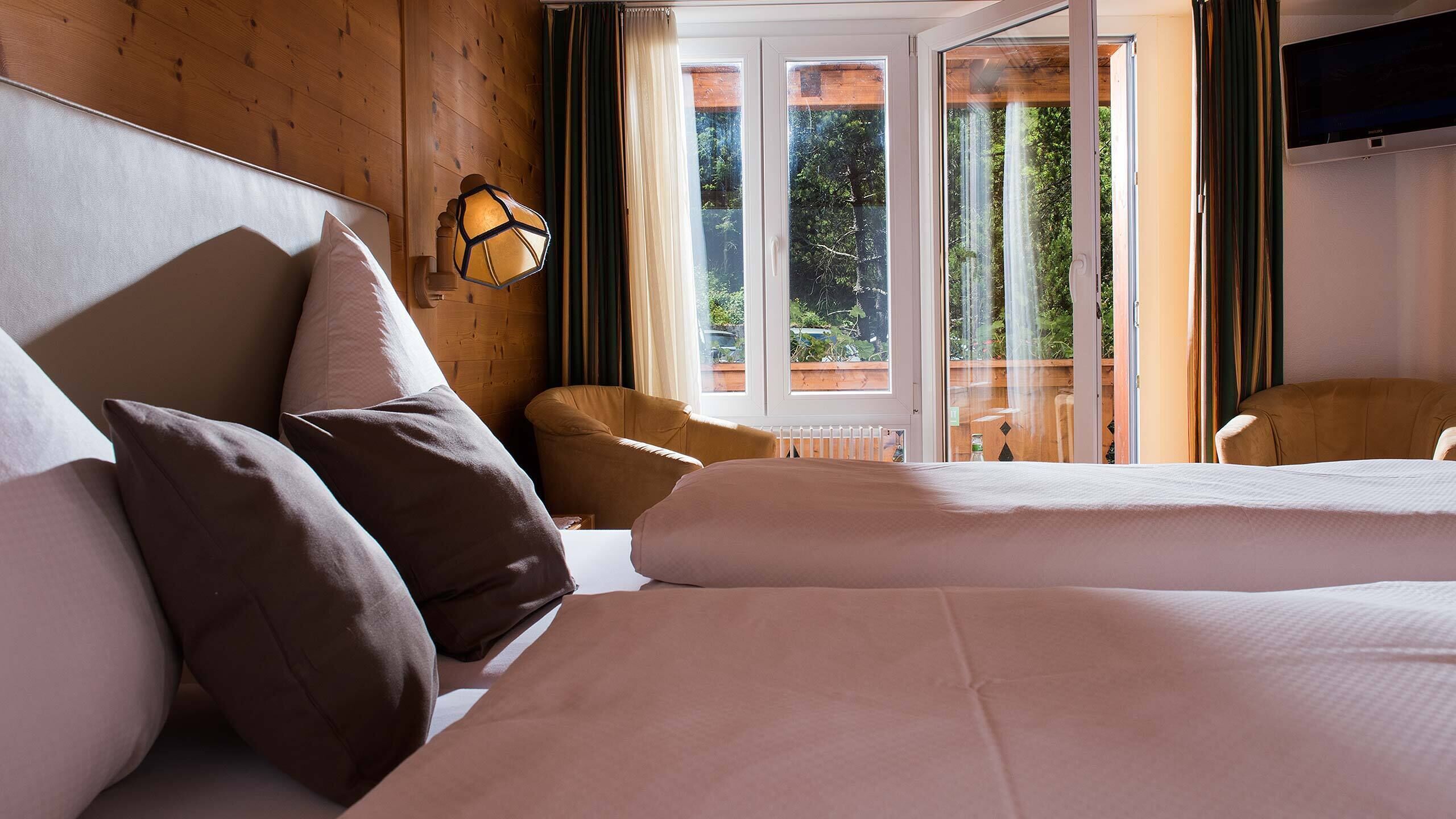 Habitación de hotel con dos camas, paneles de madera, una ventana con vista a follaje y una lámpara fijada a la pared.