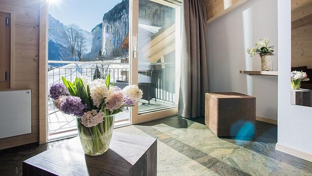 Un interior moderno con techo de madera, puerta de vidrio que da a un balcón con vistas a la montaña, un florero sobre una mesa y un taburete de madera.