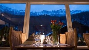 Una mesa de comedor con sillas y copas de vino, un jarrón con tulipanes rojos, y una vista de montañas nevadas a través de grandes ventanales.
