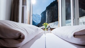 Habitación de hotel con cama, almohadas, una mesita con un florero y vistas a una montaña nevada y una cascada.