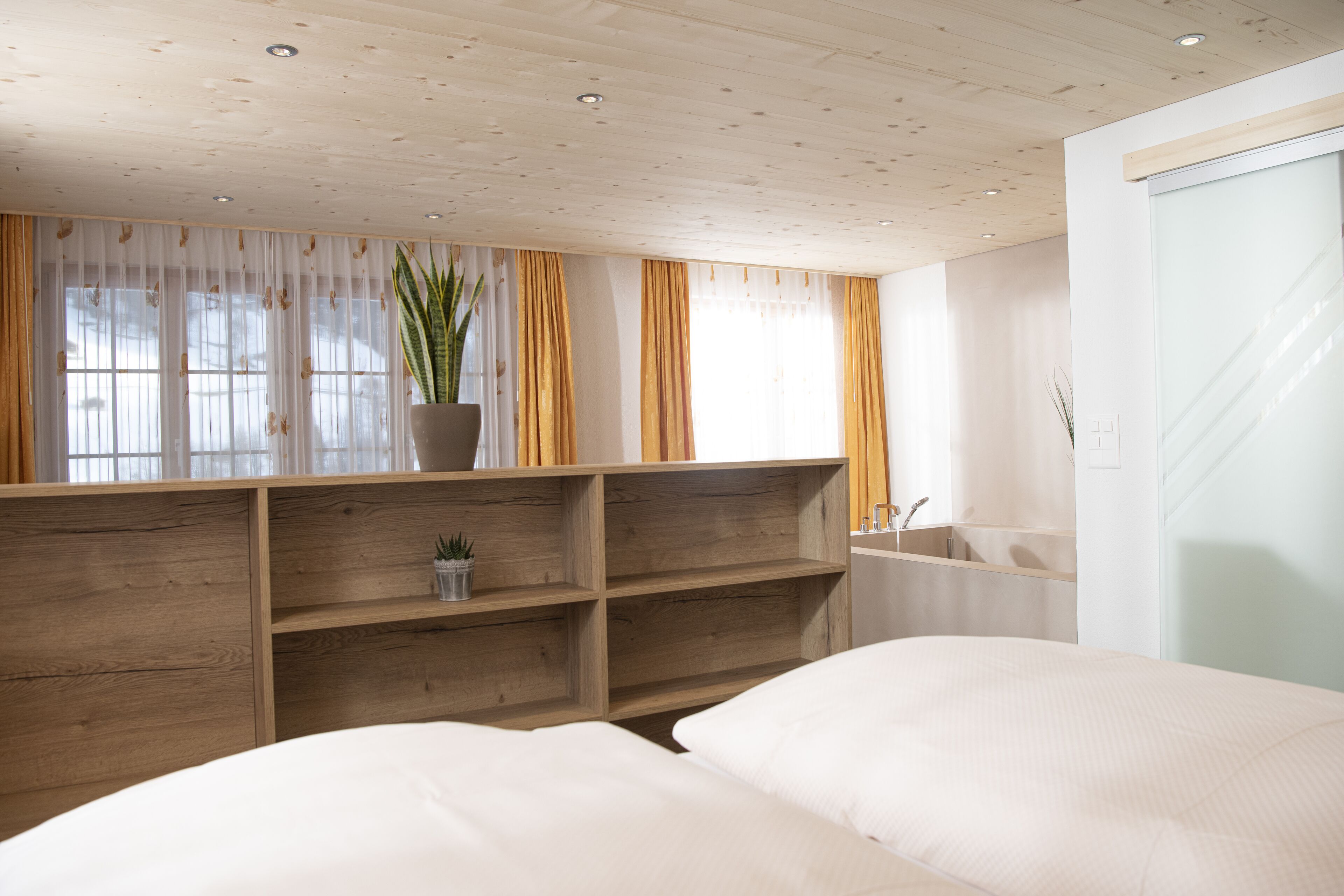 Un dormitorio con cabecera de madera, un ventanal con cortinas translúcidas, una planta en maceta y una estantería empotrada.