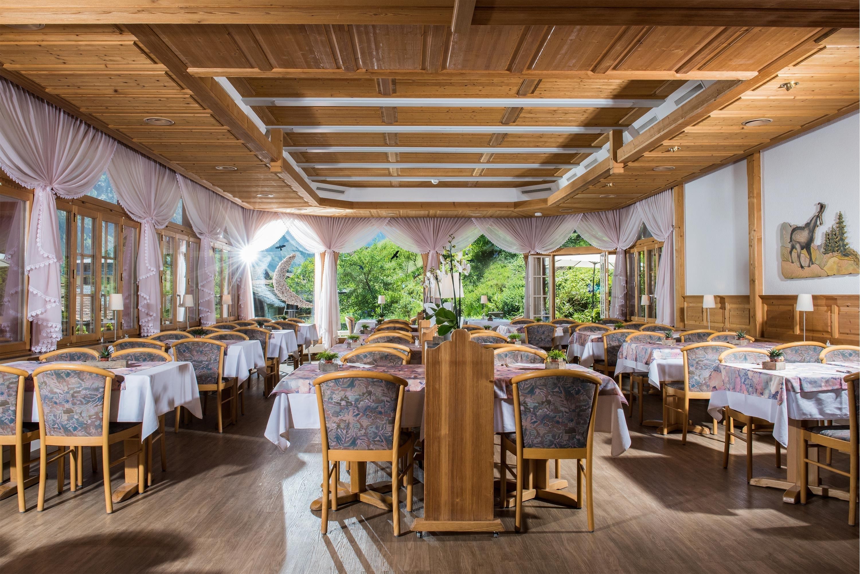 A dining area with wooden tables and chairs, large windows with curtains, and a view of greenery outside.