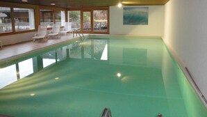 An indoor swimming pool with clear water, surrounded by windows and a wooden deck with white chairs. - Simmental (Boltigen)