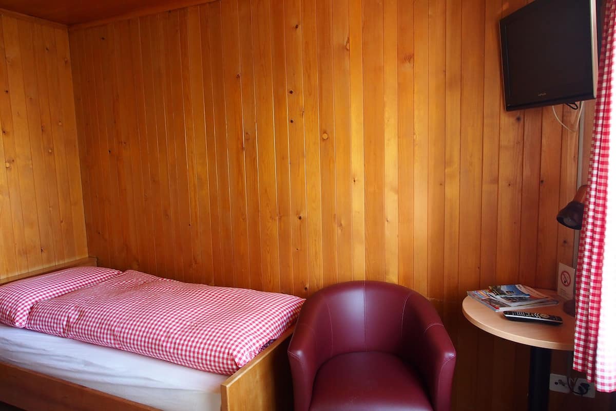 Um quarto com painéis de madeira, uma cama, uma cadeira vermelha, uma pequena mesa e uma televisão na parede.