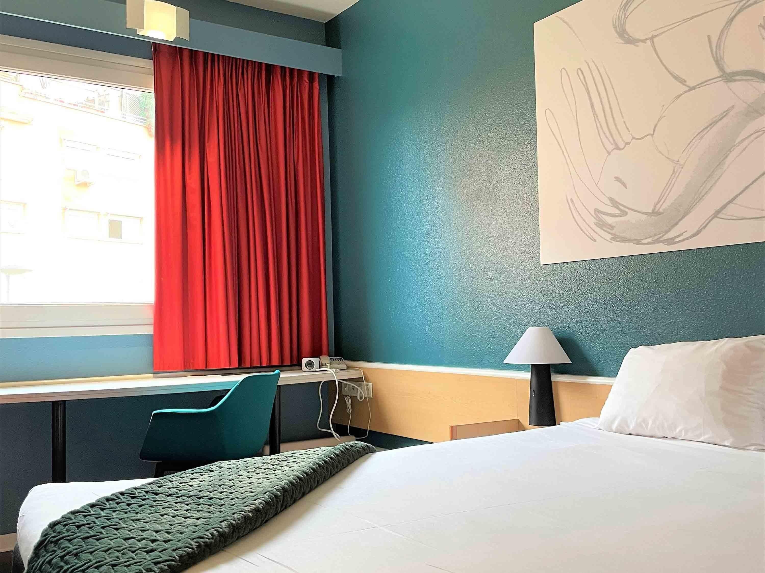 Habitación de hotel con cama, escritorio, silla, lámpara y ventana con cortinas rojas.