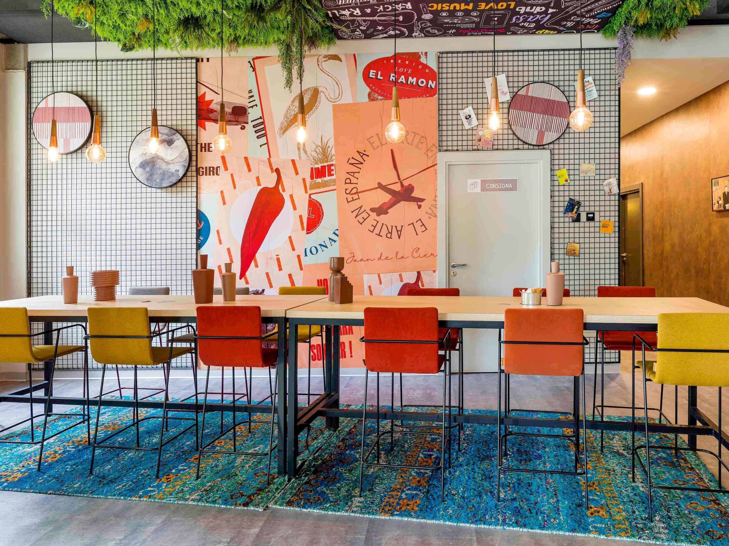 A dining area with a long table, multiple chairs, hanging lights, and a colorful rug.