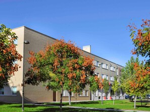 A modern building with a brick facade, surrounded by well-maintained grass and trees with orange leaves. - Hotel ibis Madrid Getafe (Getafe)