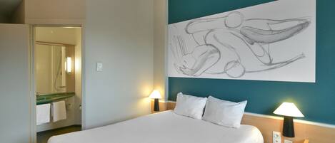Quarto de hotel com cama grande, duas luminárias de cabeceira, uma obra de arte na parede e um banheiro visível através de uma porta aberta.