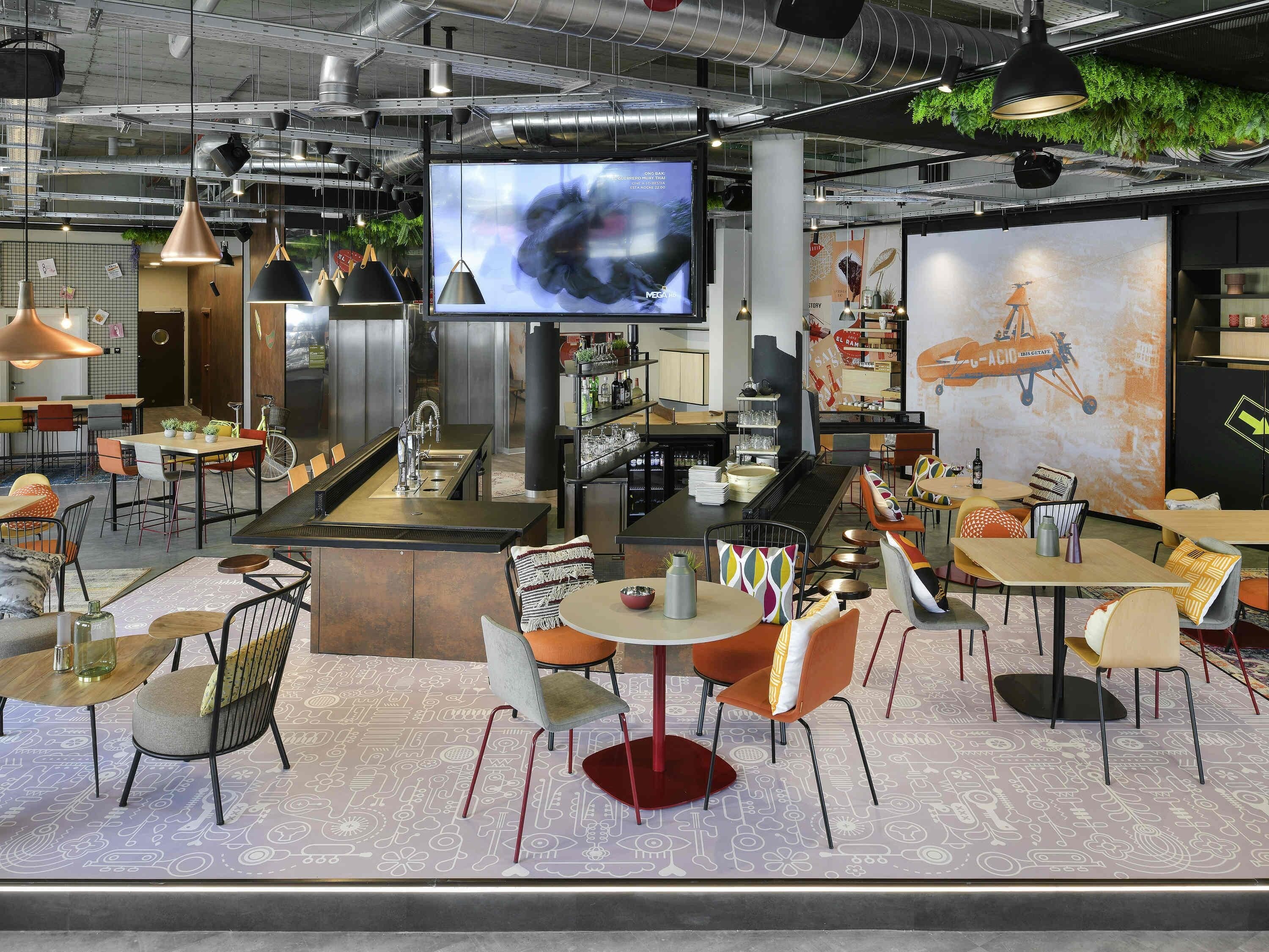 A modern cafe with a central bar, multiple seating areas, and a large screen displaying an image.