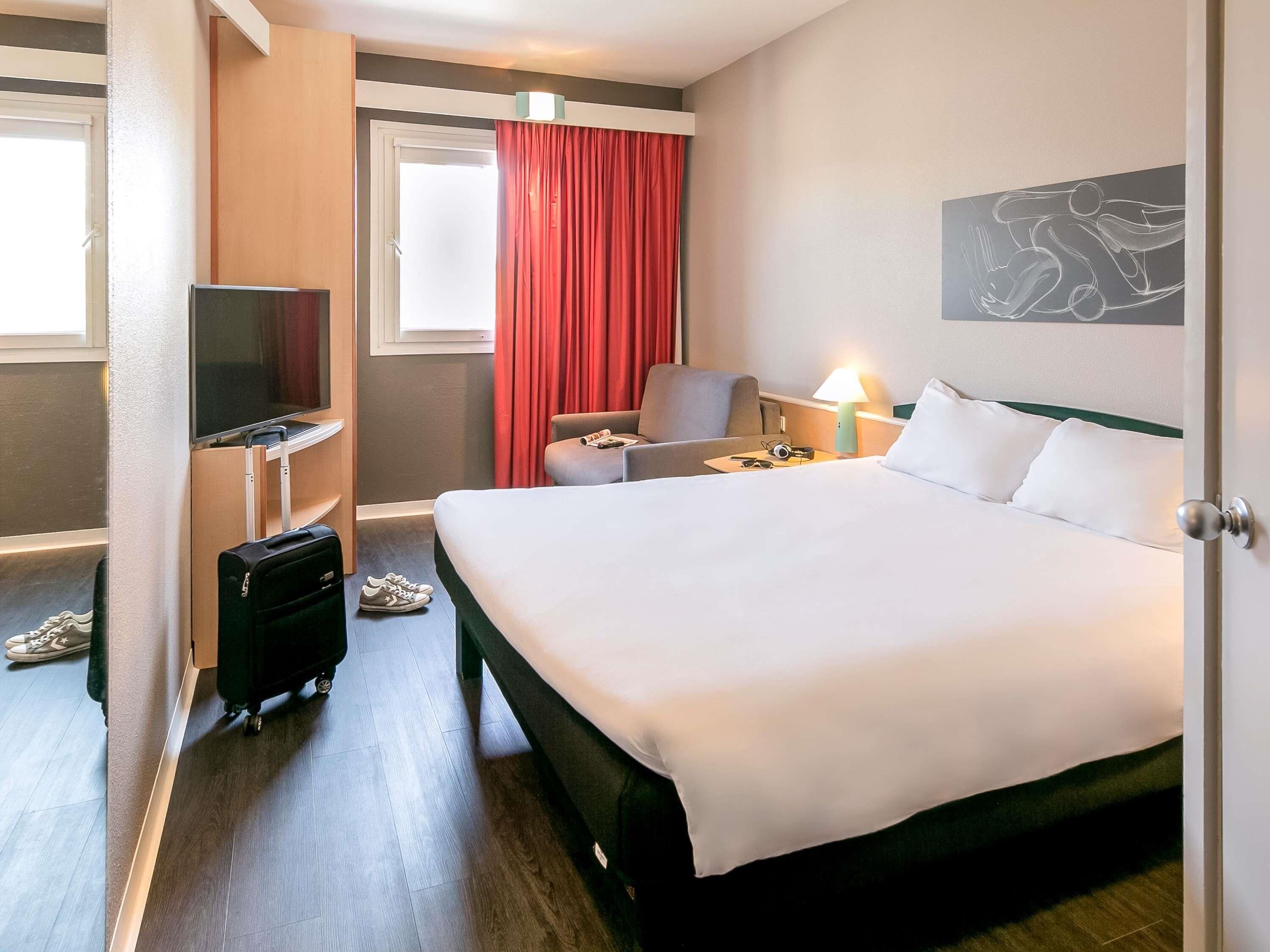 Habitación de hotel con cama, escritorio con silla, televisor y ventana con cortinas rojas.