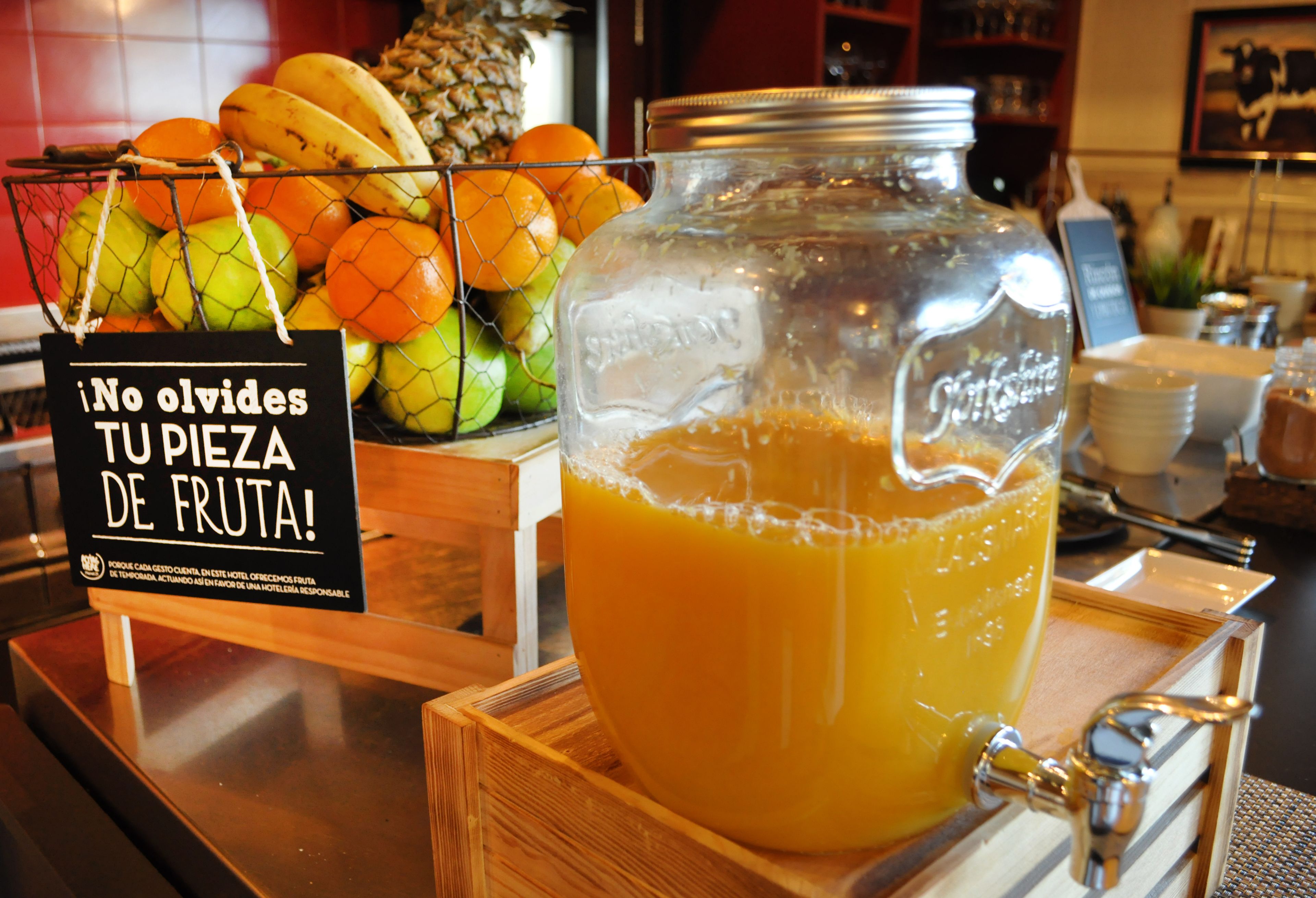 Una exhibición de frutas con un letrero, un tarro grande de jugo de naranja y una cesta de alambre con frutas.