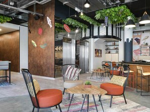 Un interior moderno con una pared decorada en madera, un comedor con mesas y sillas redondas, y una cocina con electrodomésticos de acero inoxidable.