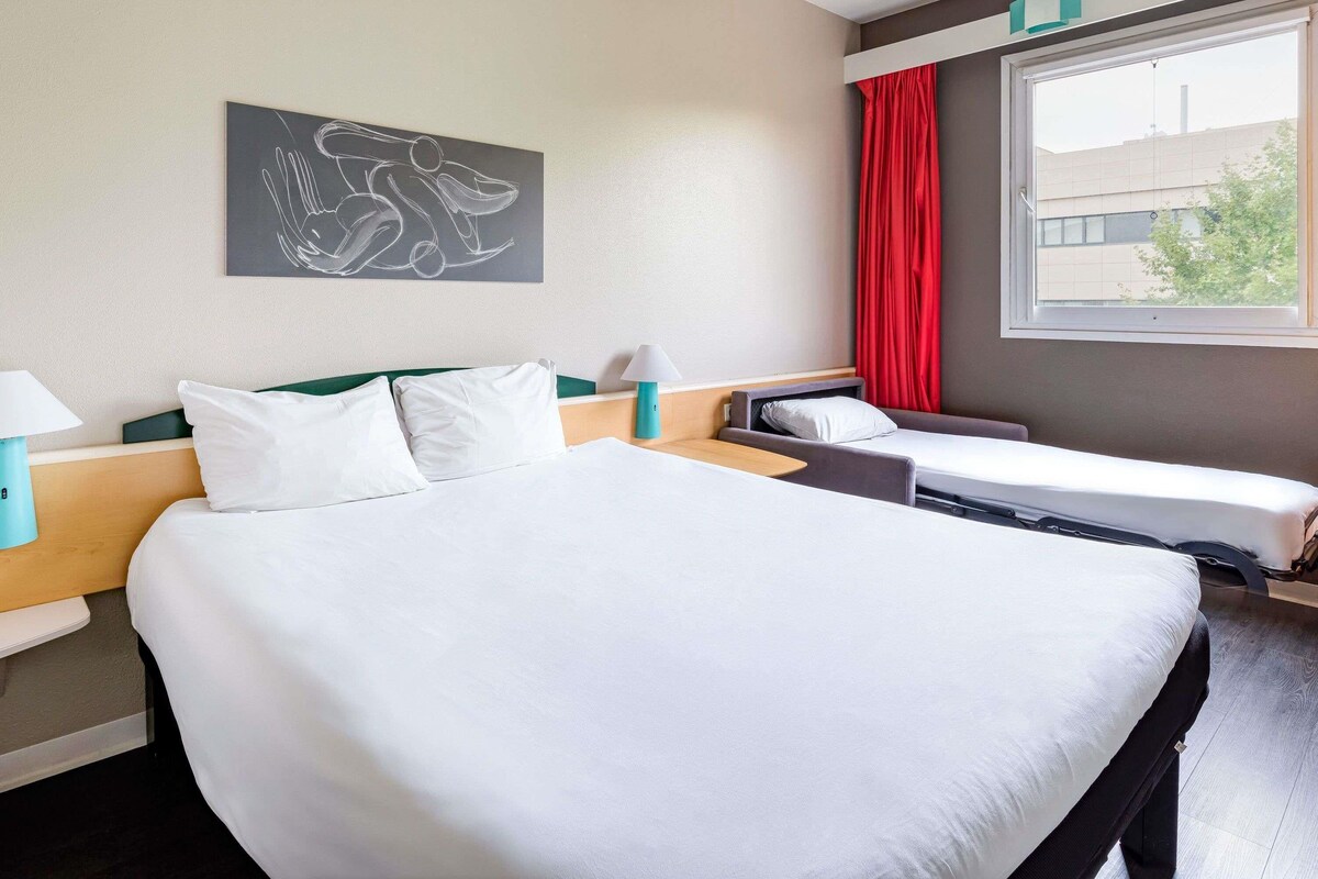 A hotel room with a large bed, two smaller beds, a window with red curtains, a wall-mounted artwork, and a lamp.
