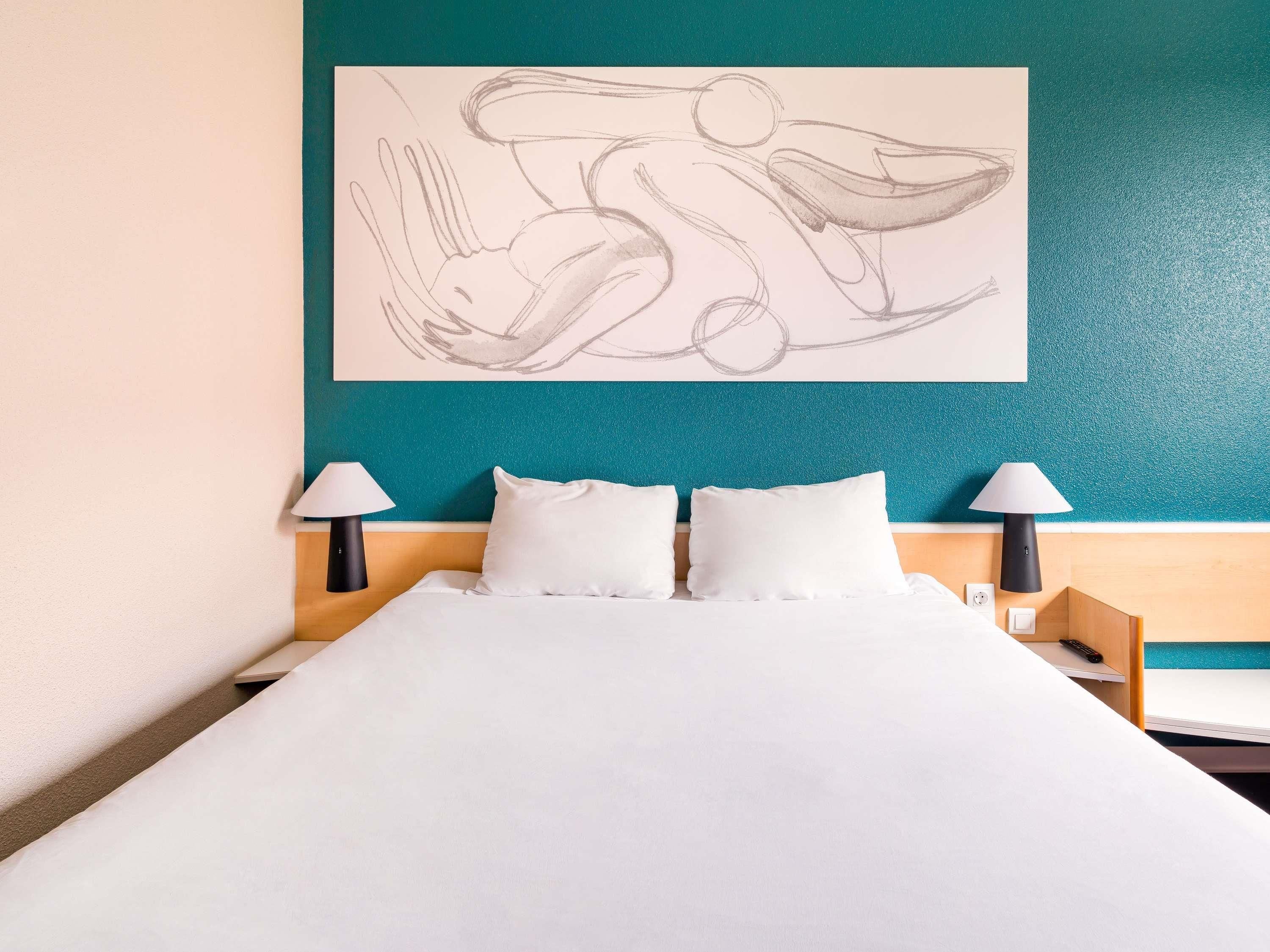 Habitación de hotel con cama, dos lámparas de noche y una pared color turquesa con un gran boceto abstracto.