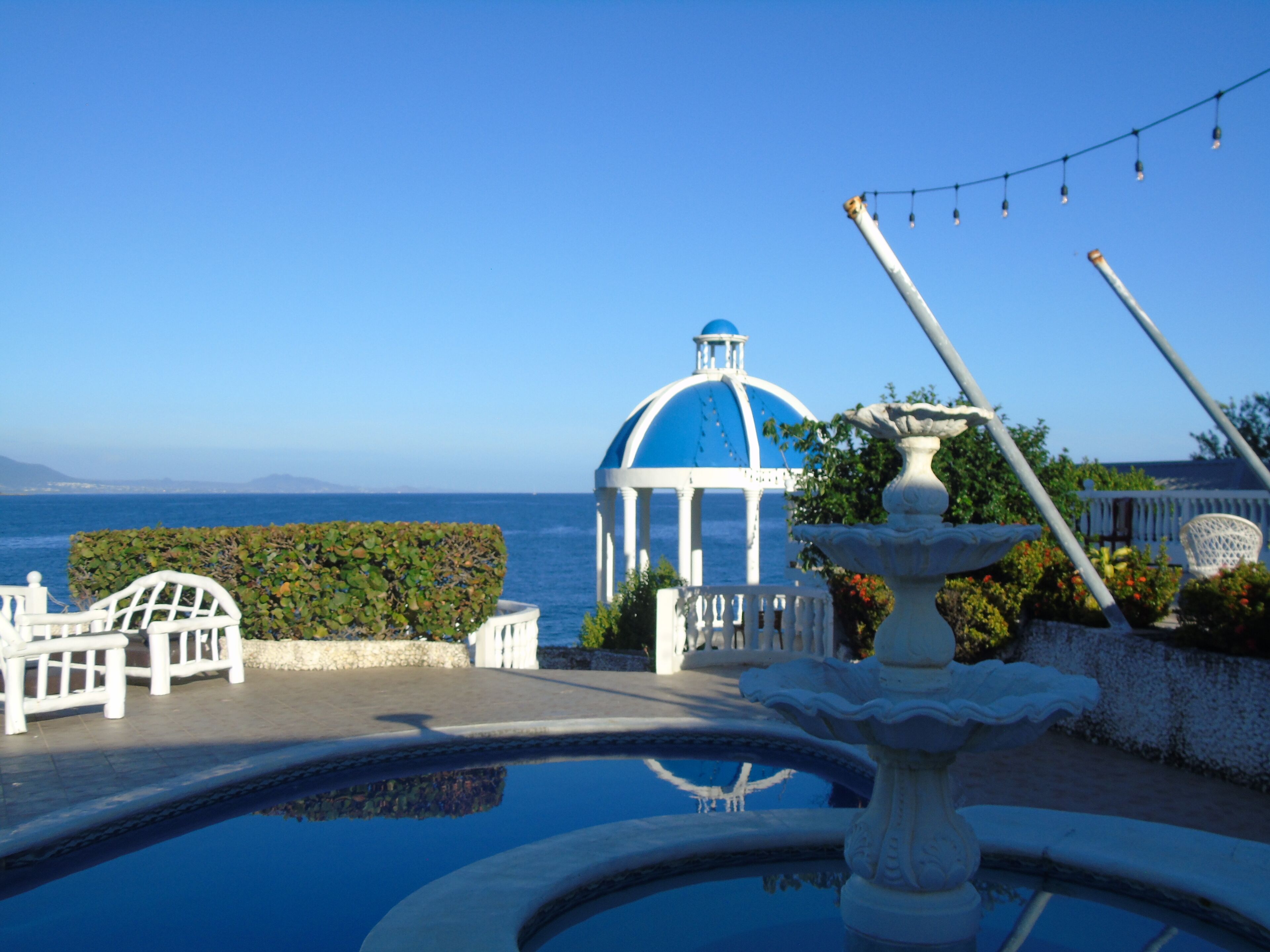 Área da piscina com uma fonte, um gazebo branco e vista para o mar.