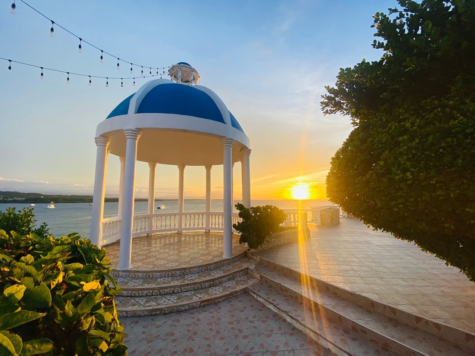 A gazebo with a blue dome and white columns, situated on a paved path with a view of the sunset over the water.
