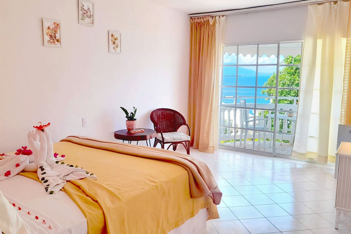 Ein Hotelzimmer mit einem Bett, einem Sessel, einem kleinen Tisch mit Pflanze und Meerblick durch ein großes Fenster.