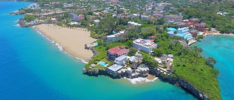 An aerial view of a coastal resort with multiple buildings, a sandy beach, and clear turquoise waters.