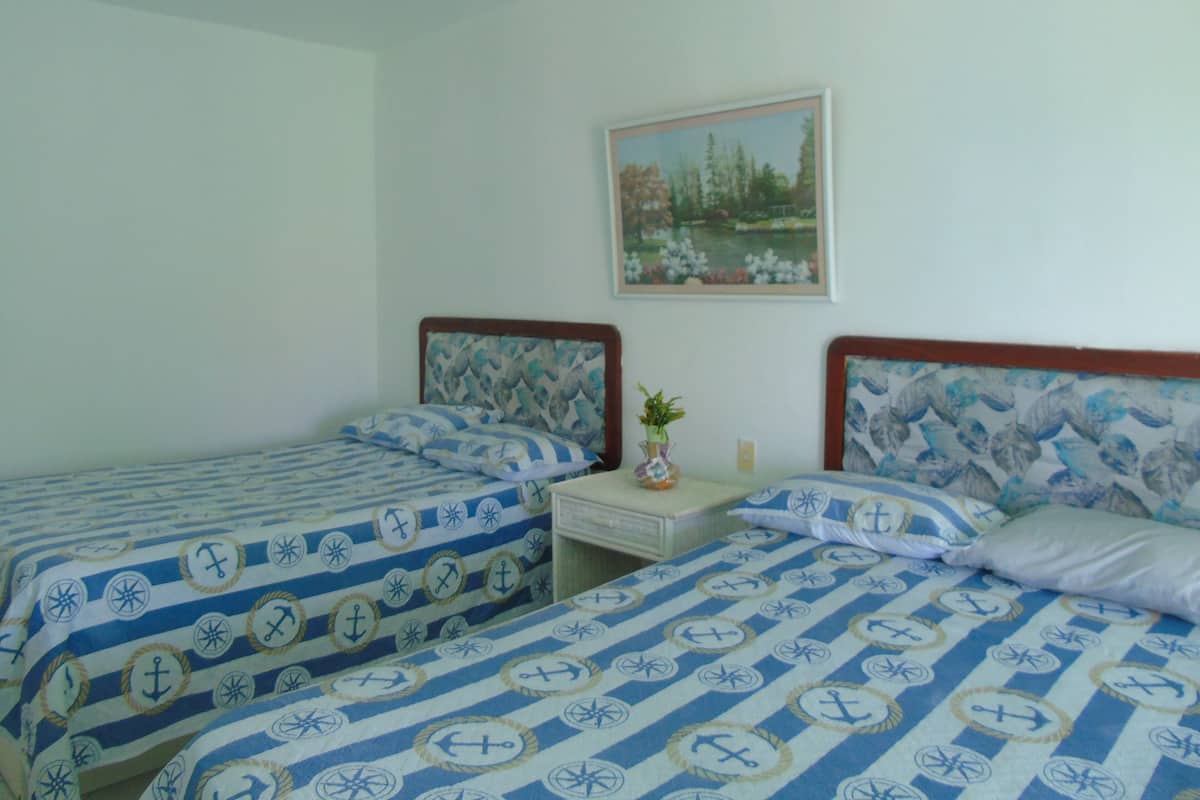 Ein Schlafzimmer mit zwei Betten, beide mit einem Bezug im Seestil. Dazwischen steht ein kleiner Nachttisch mit einer Blumenvase und einer Lampe. An der Wand über den Betten hängt ein Landschaftsbild.