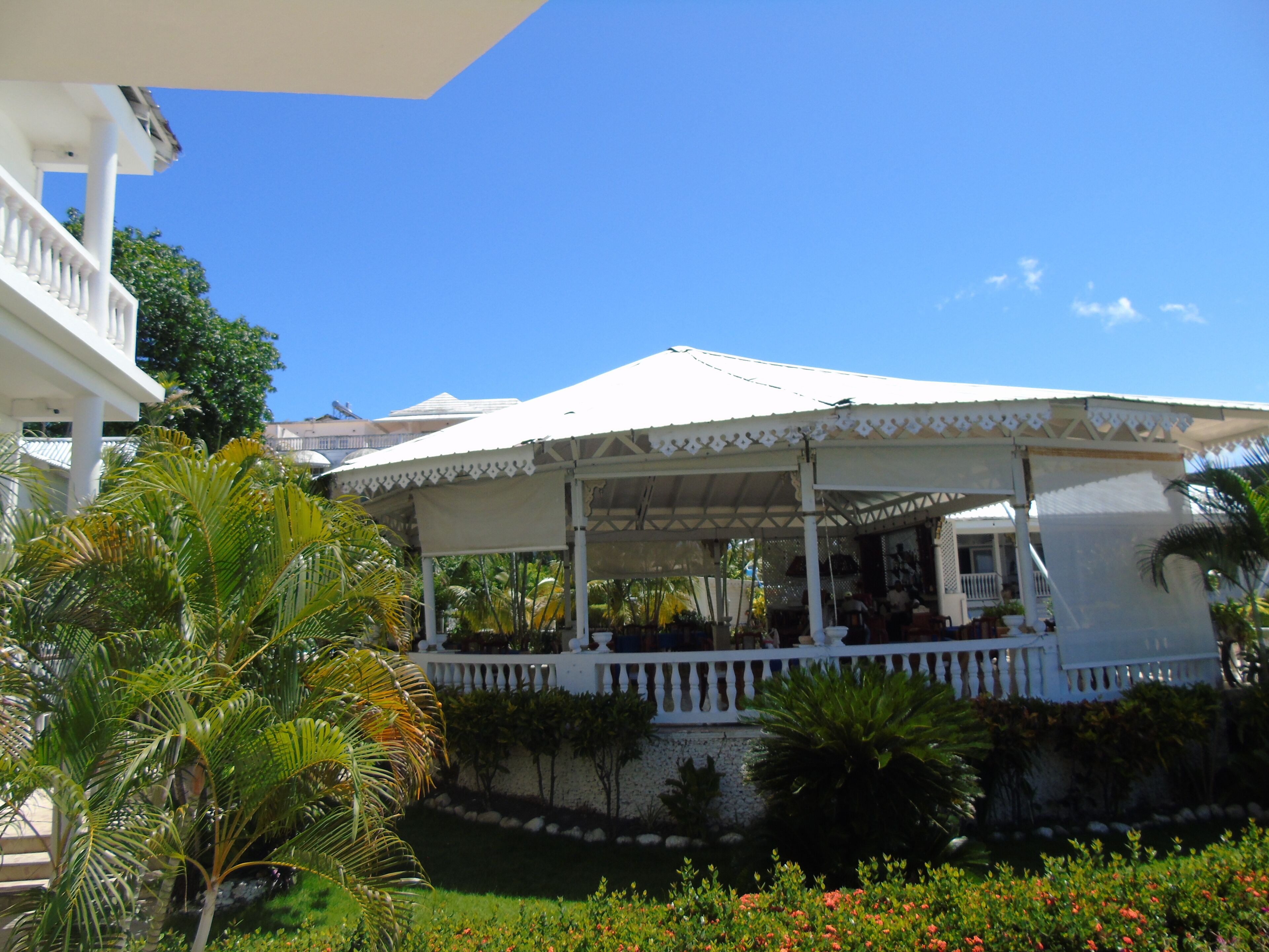 A covered outdoor area with a white railing, surrounded by greenery and a clear blue sky.