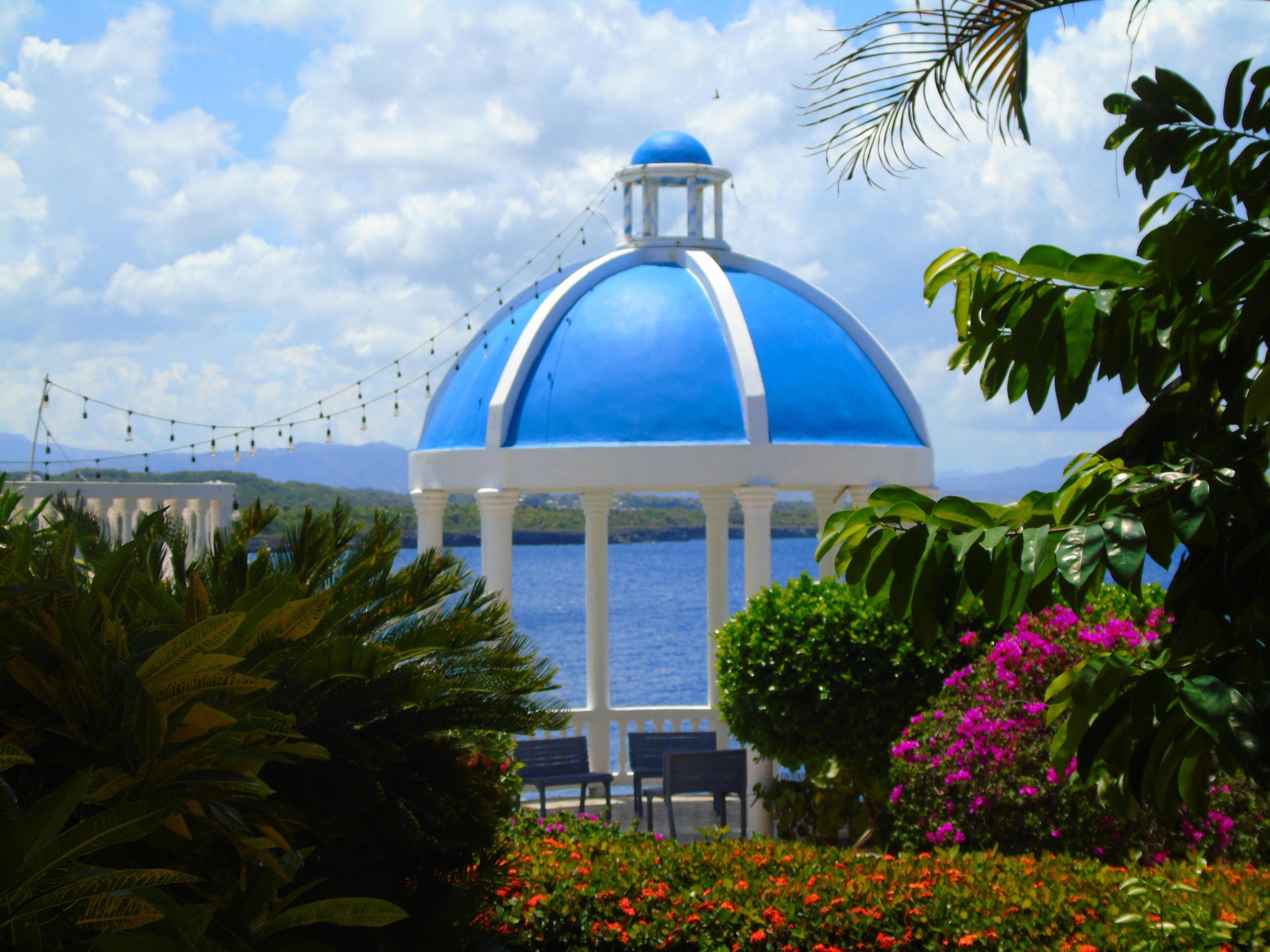 A blue-domed gazebo with white columns, surrounded by tropical plants and flowers, overlooking a body of water.