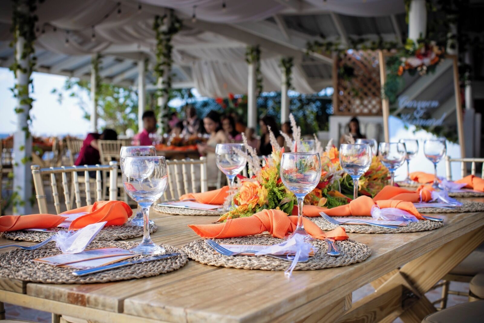 A long wooden table set with glassware, orange napkins, and woven placemats, under a tented outdoor area with greenery.