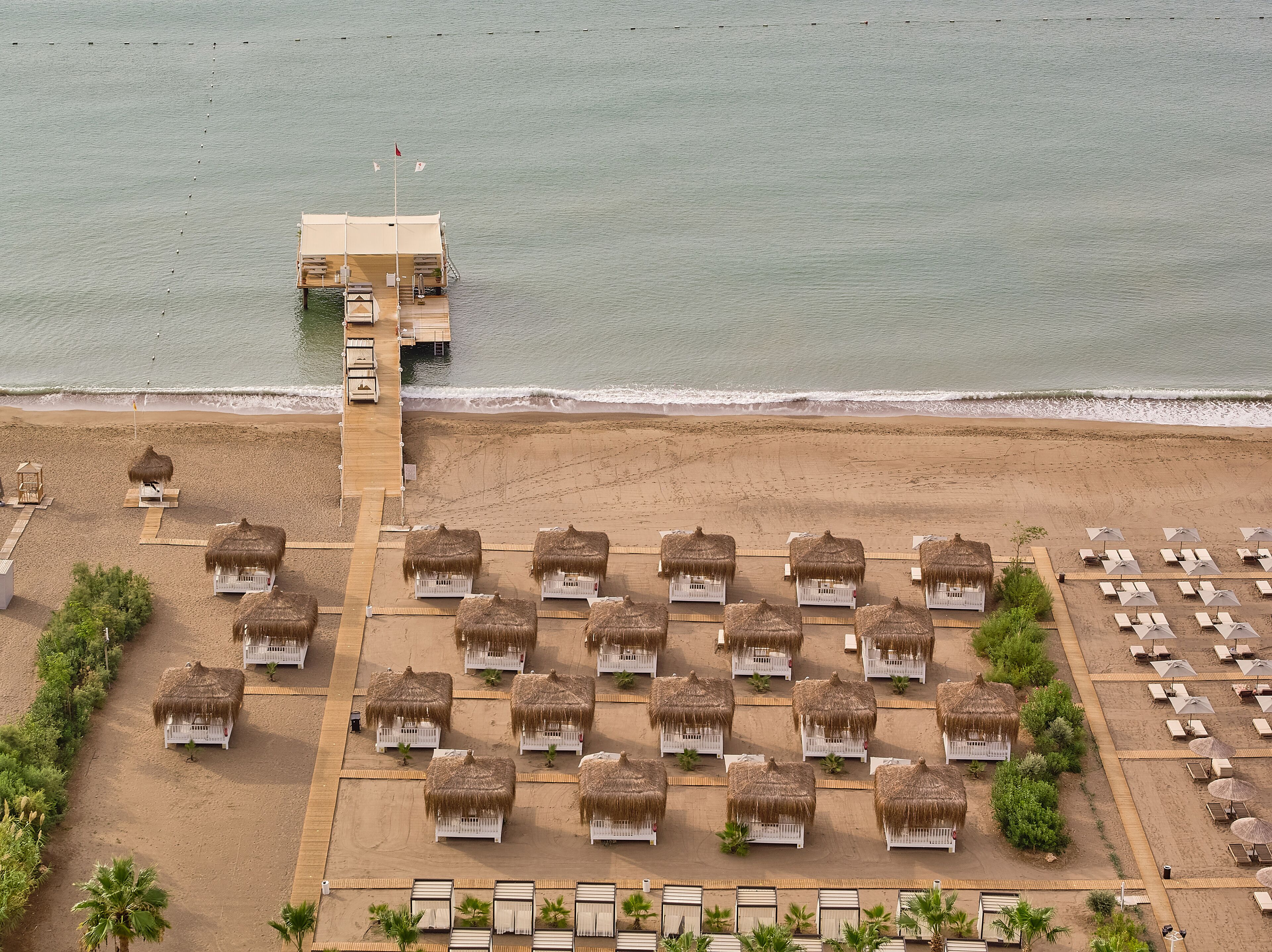 An aerial view of a beach resort with thatched-roof huts and sun loungers arranged in rows.