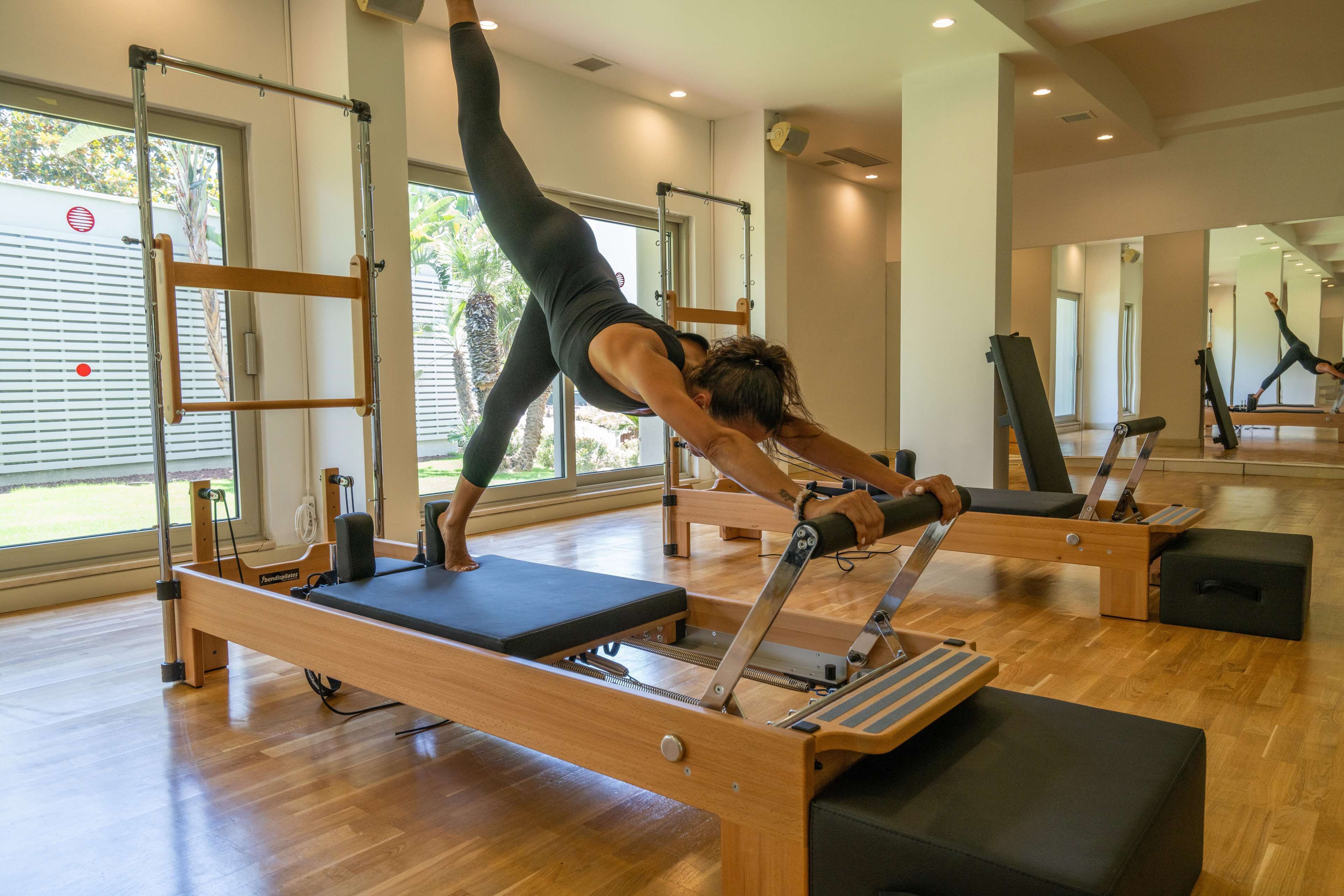 A Pilates studio with wooden floors, Pilates reformer machines, and large windows with a view of palm trees.