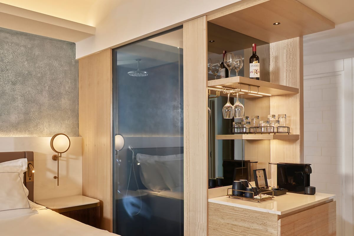 A hotel room with a bed, a bedside table, a mirror, a glass shower enclosure, a minibar, and a coffee machine.