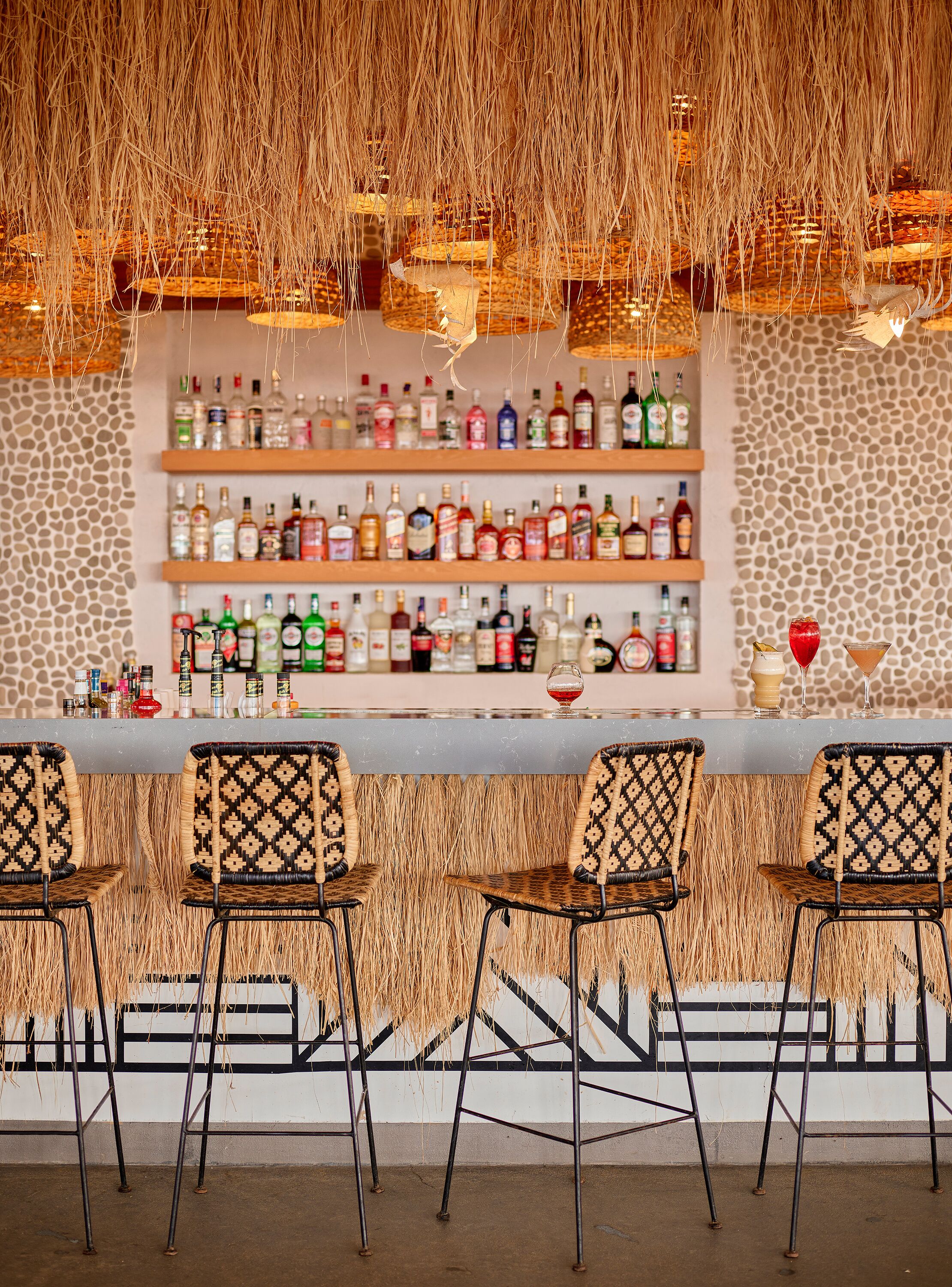 A bar area with a stone wall, shelves of liquor bottles, and woven chairs.