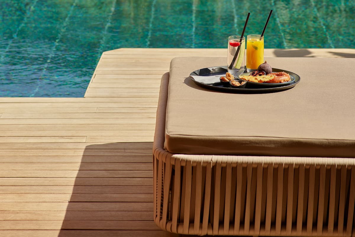 A poolside lounge area with a wooden deck, a cushioned seating area, and a dining setup with a plate of food and a glass of orange juice.