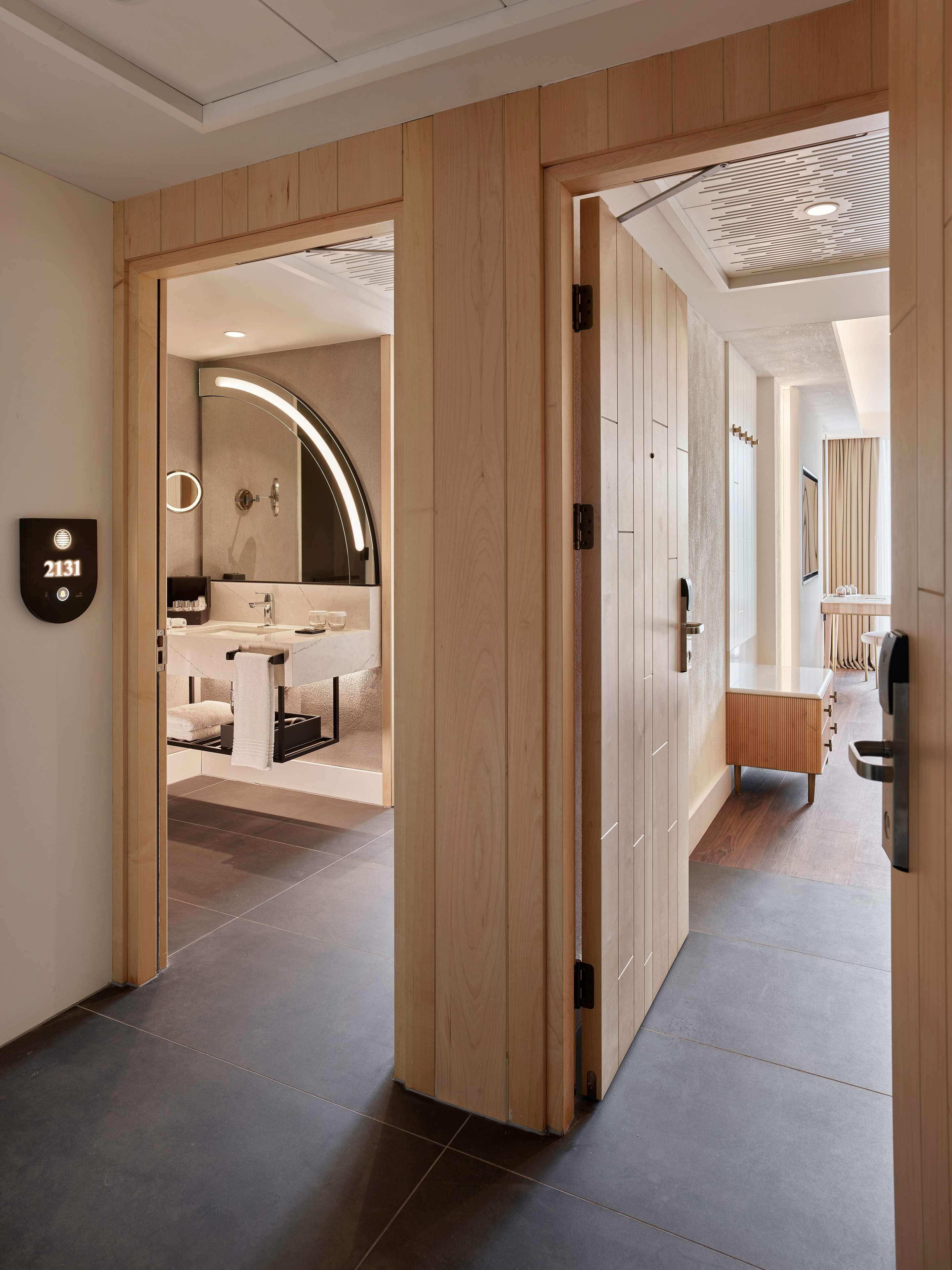 A modern hotel hallway with wooden paneling, a visible room number '2131', and a glimpse of a bathroom with a large mirror and a sink.