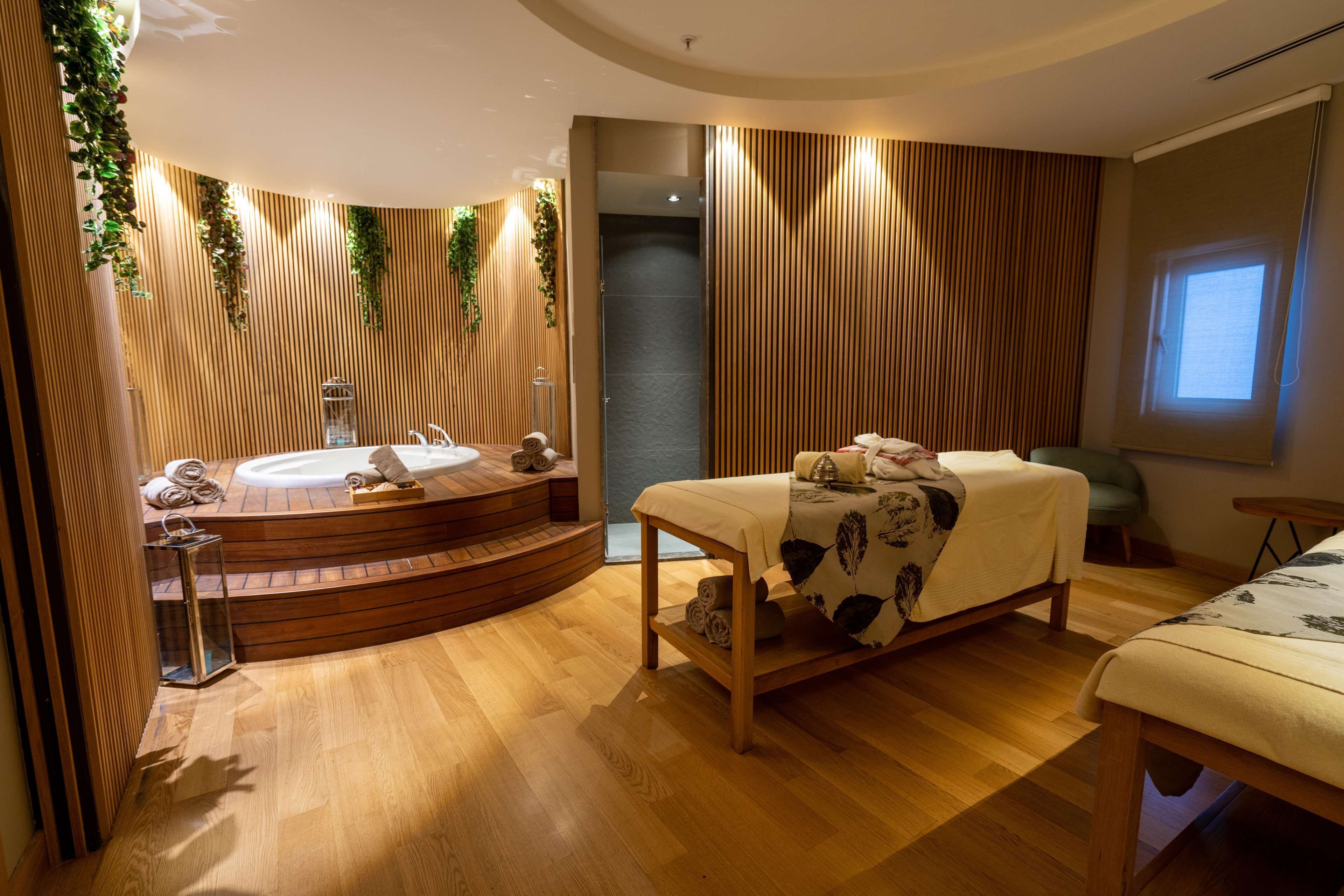 Ein moderner Spa-Raum mit Holzboden, einer Badewanne in Bogenform, einer Massageliege und einem kleinen Fenster.
