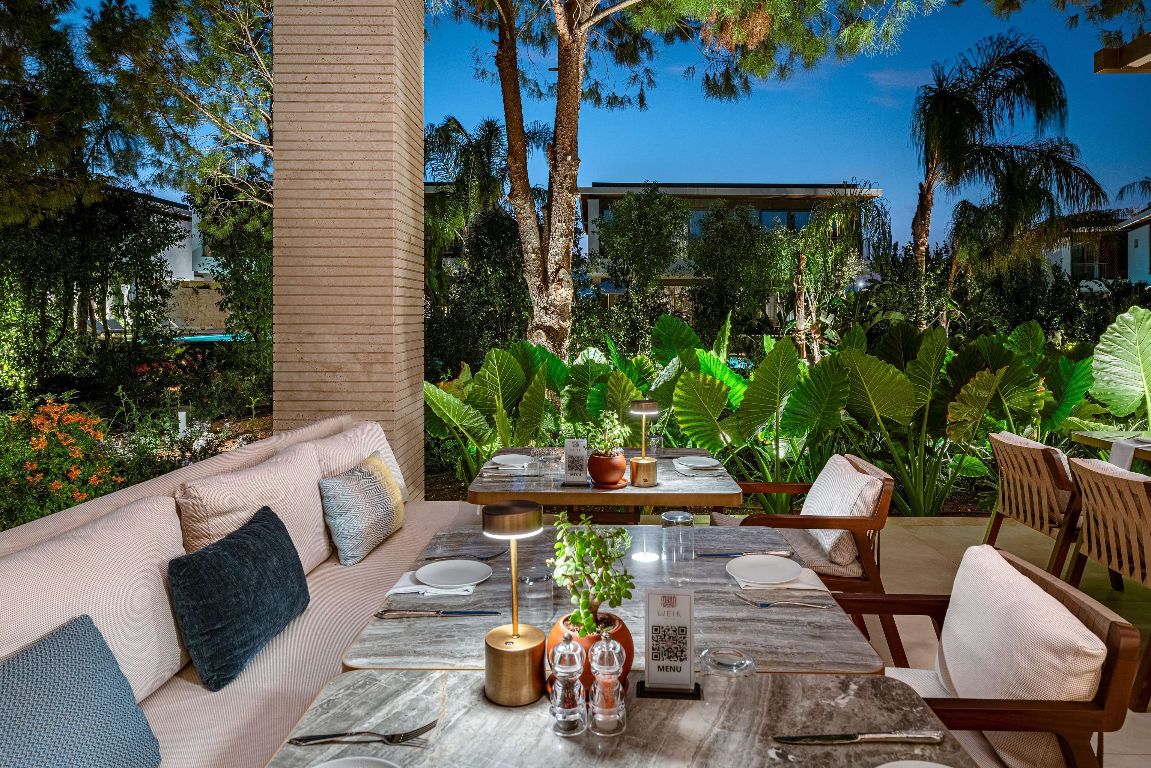Un comedor al aire libre con una mesa de madera dispuesta para dos personas, rodeado de exuberante vegetación y palmeras.