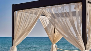 A beachside cabana with a view of the sea, a table with a drink and a magazine, and a hat on the bed.