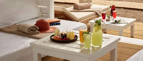 A beachside lounge with a white table set with fruit and drinks, a sofa, and a view of the ocean.