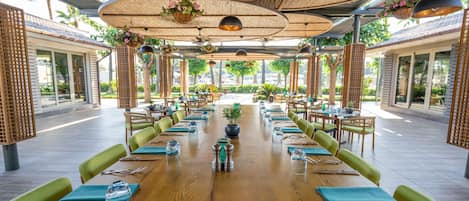 Un amplio espacio exterior para comer con mesas de madera y sillas acolchadas de color verde, rodeado de plantas y cestas de flores colgantes.