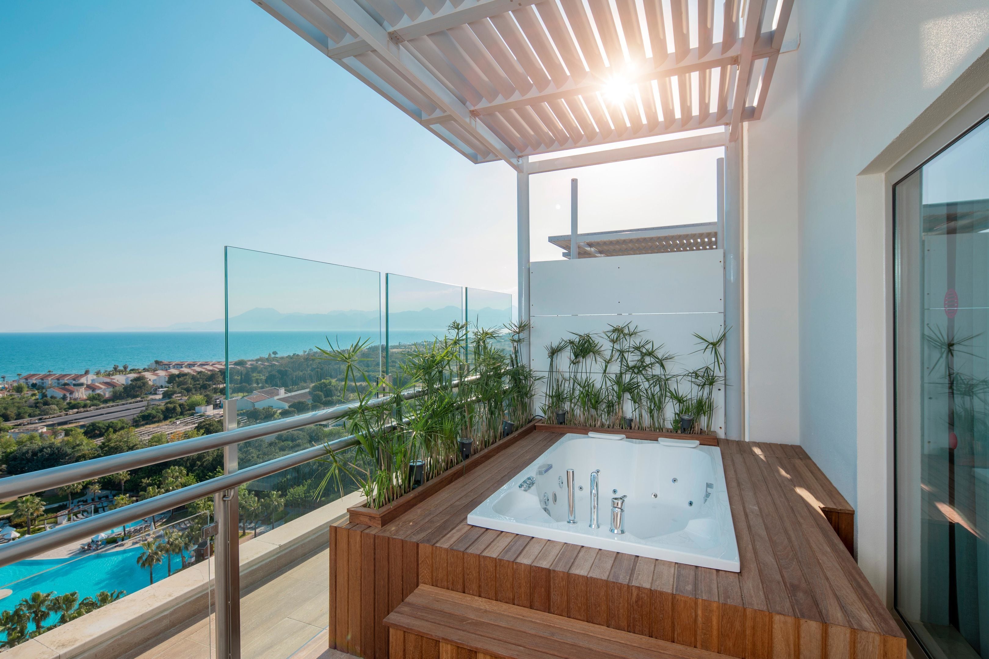 Un balcón con una bañera de madera, barandas de vidrio y vista al mar.