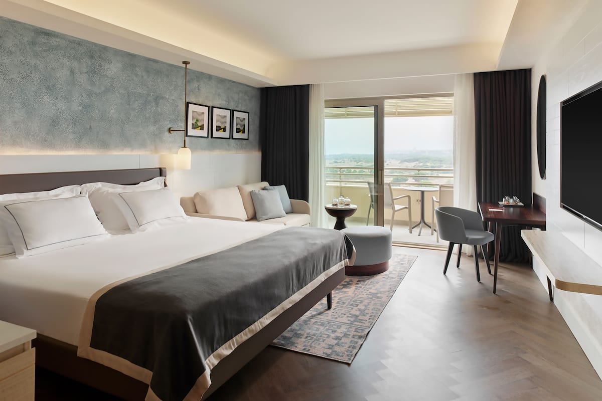A modern hotel room with a large bed, a desk, a chair, and a balcony with a view.