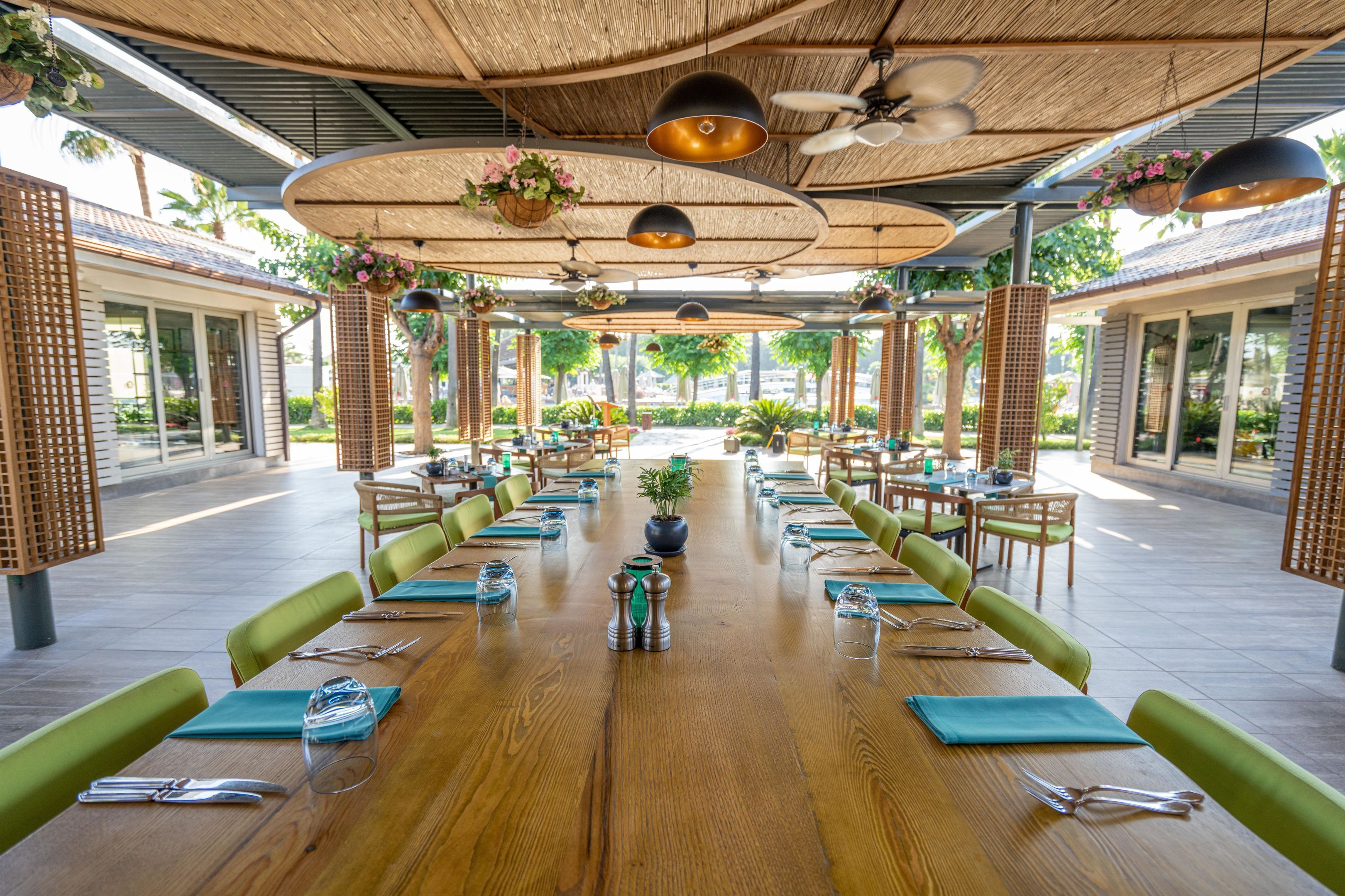 Een ruime eethoek buiten met houten tafels, groene fauteuils met kussens, hangplanten en een rieten dak.