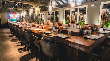A long bar with multiple stools and a counter, featuring hanging light fixtures and various bar tools.