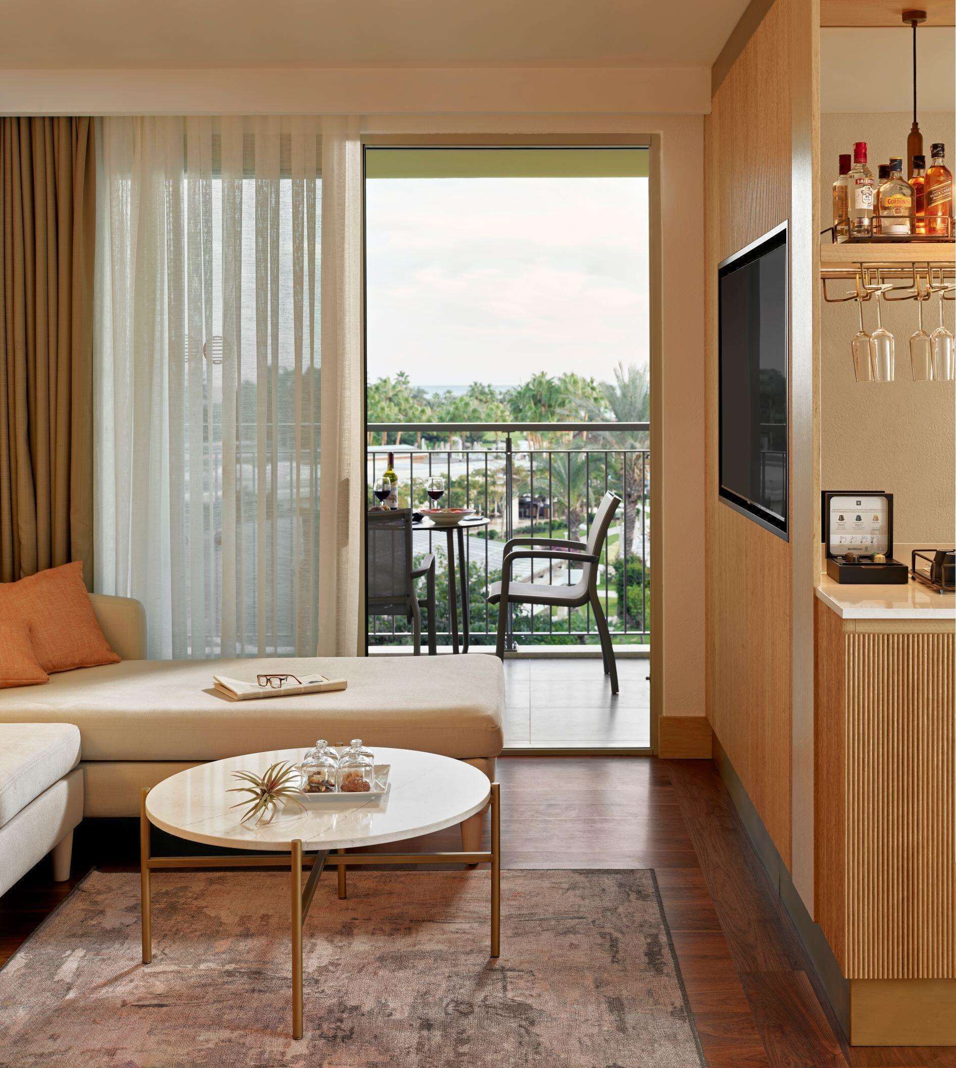 A modern hotel room with a sofa, a round coffee table, a balcony with a dining area, and a minibar.