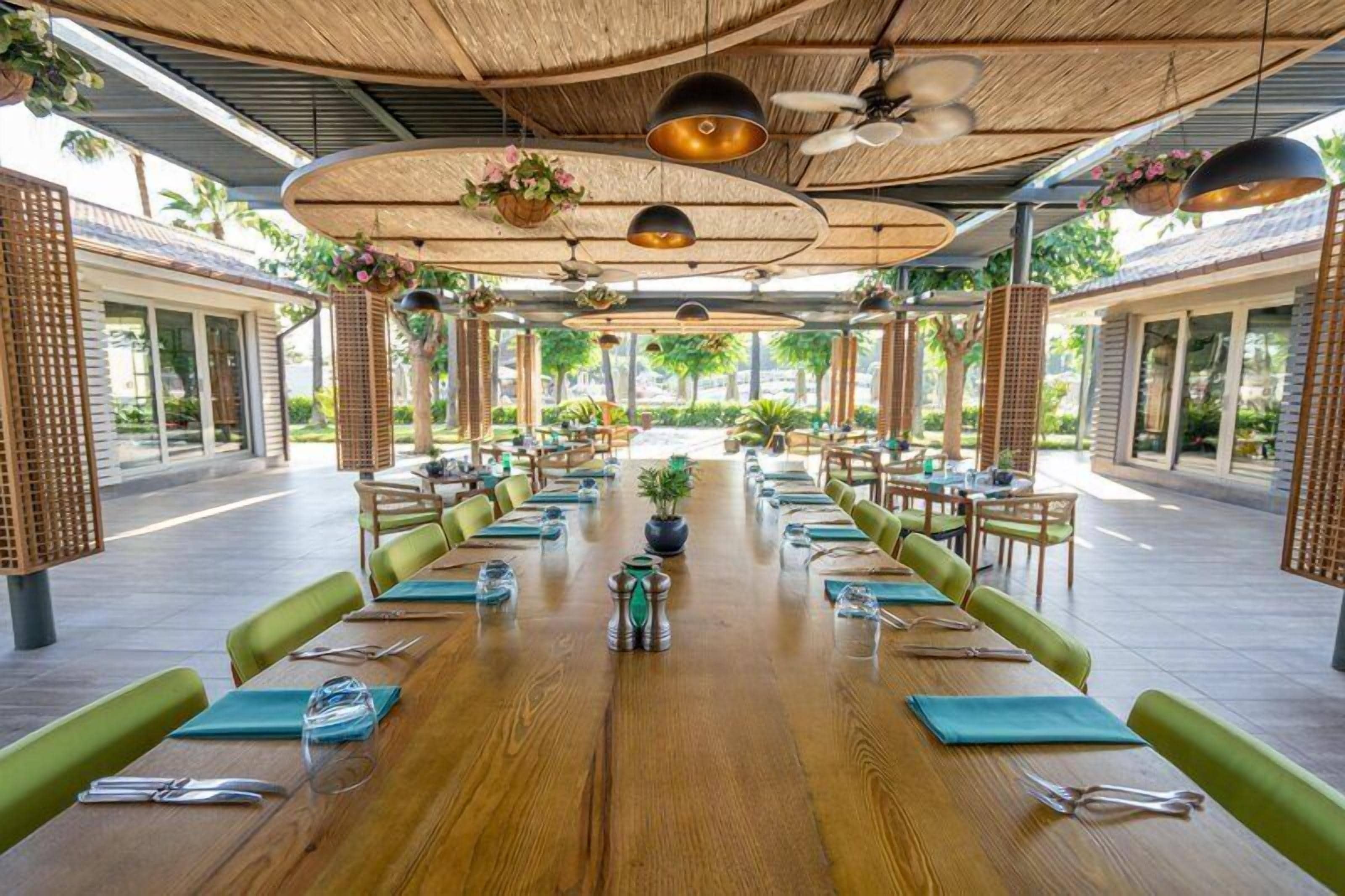 A spacious outdoor dining area with long wooden tables, green cushioned chairs, and hanging plants.